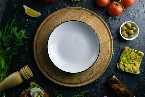 Белая тарелка с свободным местом на деревянной доске. Вокруг помидоры, суп, оливки, лимон, оливковое масло со специями и бутербродами
 - Фото, изображение