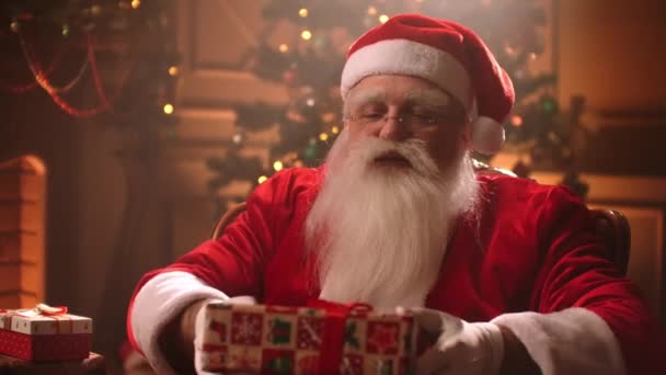 Een oudere man met een witte baard en een kerstman kostuum Handen een geschenk aan de camera en glimlacht - Video