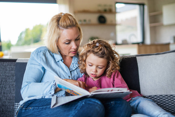 A cute small girl with mother on sofa indoors at home, reading book. - Photo, image