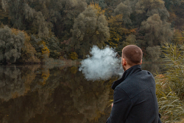 A guy with a beard sits on the lake and smokes an electronic cigarette. Blows puffs of smoke. The concept of unity with nature. Rest near the water. Vape, Man Vaping. lips and smoke, a lot of smoke. - 写真・画像