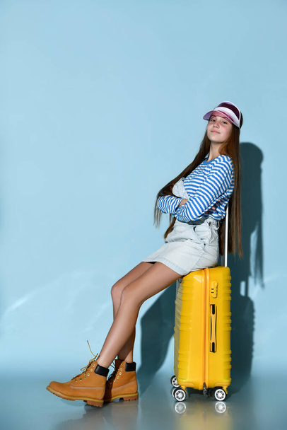 Adolescent in jeans overall skirt, sweatshirt, boots, sun visor cap. She sitting on yellow suitcase, blue background. Full length - Fotoğraf, Görsel