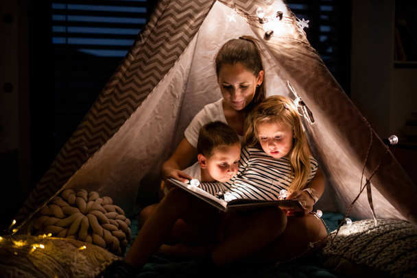 Mother with children sitting indoors in bedroom, reading a book. - Photo, Image