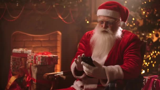 Сучасний Санта Клаус сидить на стільці на задньому плані різдвяного дерева, використовуючи смартфон, балачки і читання повідомлень від дітей.. - Кадри, відео