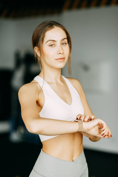 Stands after a workout with a watch. Portrait of a sports girl in the gym. Beautiful white girl for advertising.  - Фото, зображення
