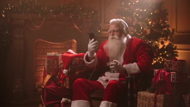 Een oudere man met een witte baard luistert naar muziek in een kerstmankostuum op kerstavond. Kerstman in het nieuwe jaar - Video