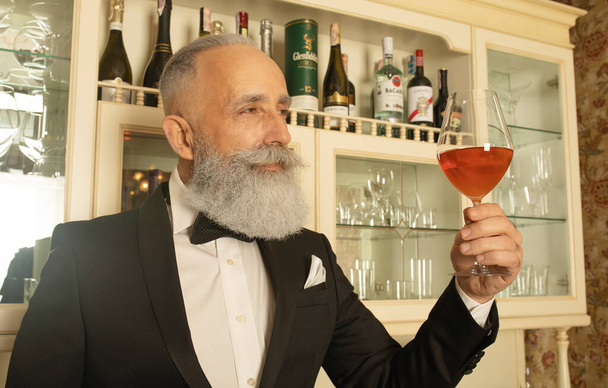 Elegant bearded senior gentleman proposing a toast with a glass of rose wine and looking at the camera. - Fotó, kép