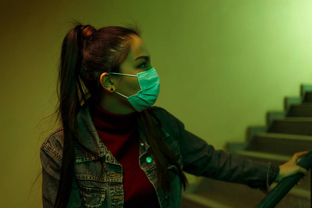 ritratto di una giovane donna asiatica in una maschera medica usa e getta. le scale dell'ospedale. la ragazza guarda indietro con ansia. paura
 - Foto, immagini