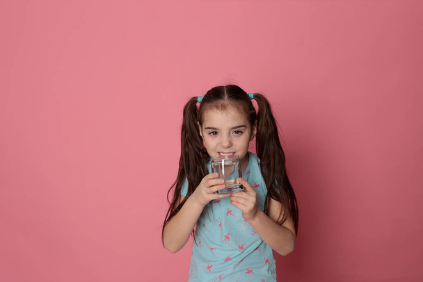 Una bella ragazza di otto anni beve acqua da un vetro trasparente su sfondo rosa in una t-shirt blu
 - Foto, immagini