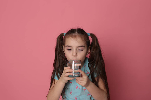 Una bella ragazza di otto anni beve acqua da un vetro trasparente su sfondo rosa in una t-shirt blu
 - Foto, immagini