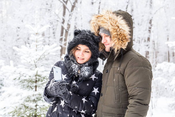 テクノロジーと関係の概念-幸せな笑顔のカップルは外の冬の森の中で自撮り - 写真・画像