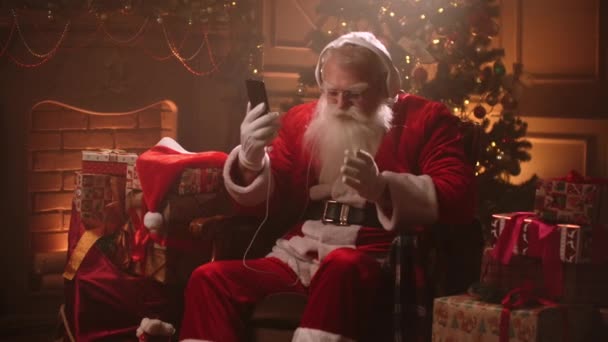 Tańczący Święty Mikołaj ze słuchawkami i telefonem komórkowym w rękach. Słuchaj muzyki online. Jerky i dynamiczne piosenki - Materiał filmowy, wideo