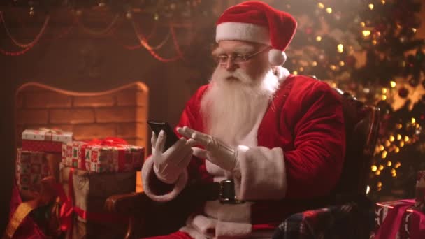 De Kerstman zit in een stoel op de achtergrond van een kerstboom met behulp van een smartphone en chatten op het internet en berichten lezen van kinderen. - Video