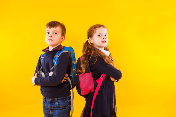 Schoolgirl and schoolboy with backpacks and crossed hands standing over yellow background - Fotoğraf, Görsel