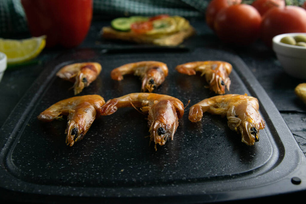 Grey cutting board with shrimp. Around tomato, souce, olives, lemon, olive oil with spice and sandwiches - Valokuva, kuva