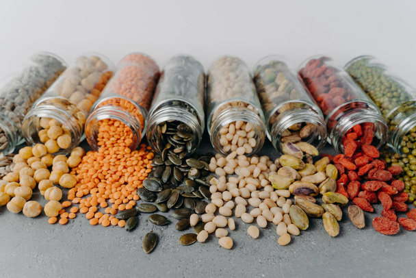 Colpo orizzontale di vari semi proteici, fagioli e cereali versati dalle bottiglie di vetro sulla superficie grigia. Prodotti crudi per gli amanti della salute
 - Foto, immagini