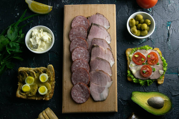 Sliced salami and ham on a wooden board, around the olives board with other sandwiches, tomato, lemon and sauce. - Photo, Image