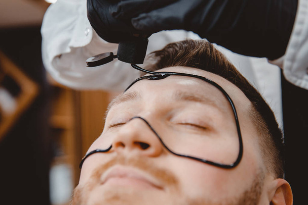 Barber applique un masque au charbon noir sur le visage de l'homme pour nettoyer la peau des pores et enlever l'acné du nez, salon de spa
 - Photo, image