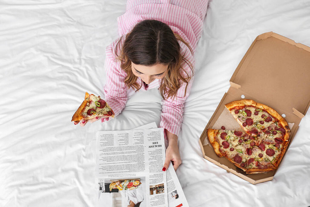Morgen der schönen jungen Frau, die leckere Pizza isst und Zeitung im Schlafzimmer liest - Foto, Bild