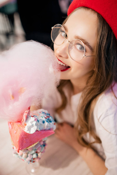 Young woman in red beret and glasses has sweet dessert with candy floss, milkshake, dumplings and pink chewing gum cake in France cafe - Φωτογραφία, εικόνα