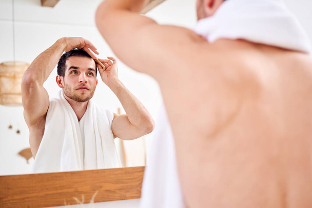 Male combing his hair while standing with white towel on neck near mirror in bathroom - Fotó, kép