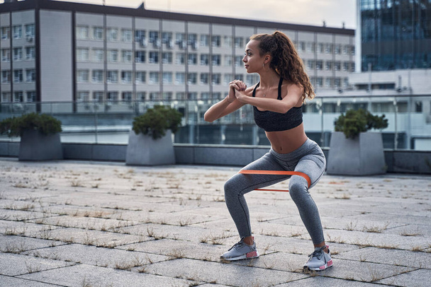 Молодая спортсменка делает приседания упражнения со спортивной резиной в городской местности
 - Фото, изображение