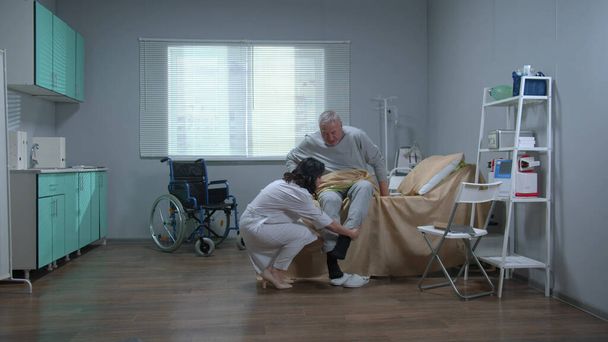 Врач в белой форме помогает старому пациенту встать с кровати
 - Фото, изображение