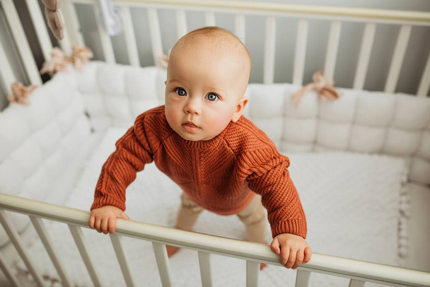 Beautiful little girl in a crib in a cozy light room in a sweater of fashionable shades. learns to stand at the support. - Fotografie, Obrázek
