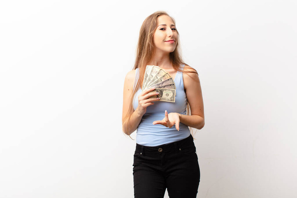 yound blonde woman smiling happily with friendly, confident, positive look, offering and showing an object or concept holding dollar banknotes - Foto, immagini