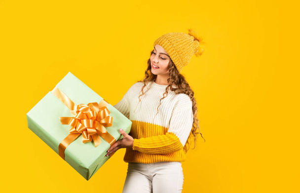 she love Christmas. xmas gift shopping sale. merry christmas to you. winter holiday discounts. joyful little girl knitted hat and sweater. kid hold present box yellow background. happy new year - 写真・画像