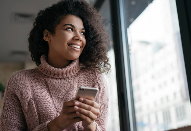 Fiatal, érzelmes afro-amerikai nő, mobiltelefonnal a kezében, online vásárlás, ételrendelés. Portré gyönyörű lány segítségével smartphone alkalmazás, nézi az ablakot, és mosolyog, ül a kávézóban - Fotó, kép