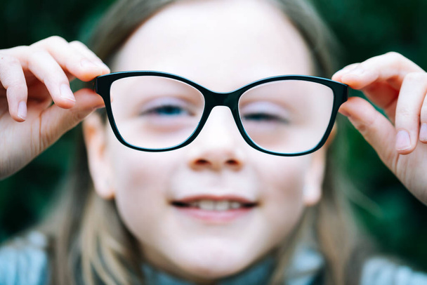 Nahaufnahme Porträt eines kleinen Mädchens mit kurzsichtiger Korrekturbrille. Mädchen hält ihre Brille mit zwei Händen direkt vor der Kamera - Brille im Fokus - geringe Schärfentiefe - Foto, Bild