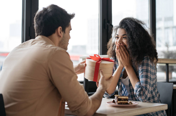Romantic man giving birthday gift to beautiful emotional woman. Portrait of lovely multiracial couple sitting together in cafe, dating. Valentines day concept - Foto, immagini
