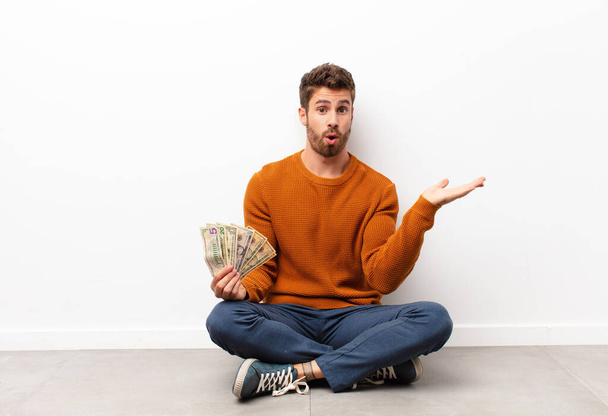 young handsome man looking surprised and shocked, with jaw dropped holding an object with an open hand on the side holding dollar banknotes - 写真・画像