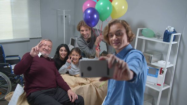 Familieleden bezoeken meisje die op het bed liggen in het ziekenhuis, jongen maakt een grappige groepsfoto - Foto, afbeelding