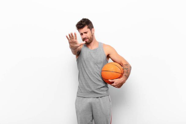 young handsome man covering face with hand and putting other hand up front to stop camera, refusing photos or pictures holding a basketball ball. - Φωτογραφία, εικόνα