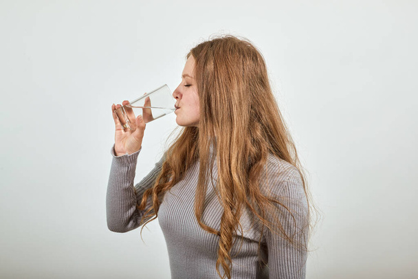 red haired woman in gray sweater quenches her thirst and drinks glass of water - Foto, afbeelding