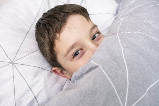 Smiling preteen boy child lying in bed and covering his tricky playful face with a blanket, pranking kid concept - Foto, Bild