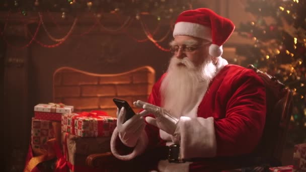 De Kerstman zit in een stoel op de achtergrond van een kerstboom met een smartphone en bekijkt zijn online bankrekeningen. - Video