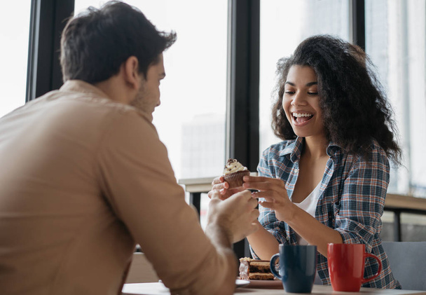 Young lovely couple laughing, sitting in cafe. Beautiful emotional African American woman eating tasty cake. Happy multiracial friends communication together. First date concept - Foto, immagini