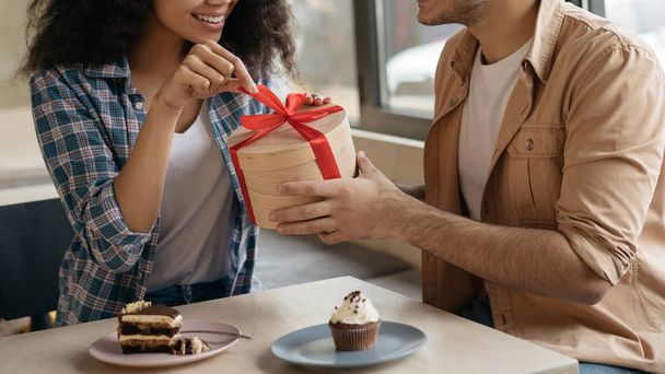 Beautiful African American woman opening gift dox with red ribbon. Lovely couple celebration Valentines day, sitting together in cafe and laughing. Romanic date concept  - Фото, изображение