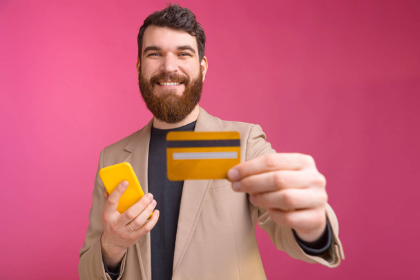 Retrato de un joven sonriente en traje que sostiene el teléfono inteligente y muestra su tarjeta de crédito
 - Foto, Imagen