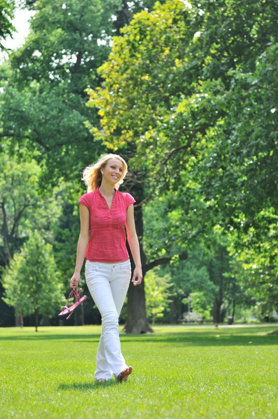 Улыбающаяся молодая женщина держит туфли и ходит босиком по траве в парке
                    - Фото, изображение