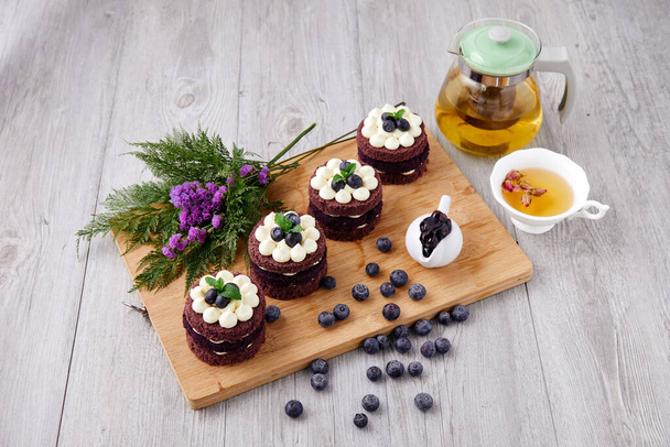 Řada čtyř hnědých dortů s modrými bobulemi na vrcholu, které společně porušují květinu, čajový hrnec a bobule jako zdobený předmět na dřevěném talíři - Fotografie, Obrázek