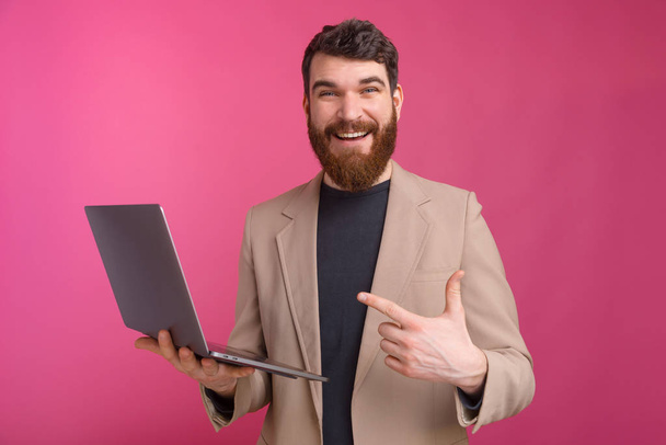Веселый молодой человек стоит на розовом фоне и указывает на ноутбук
 - Фото, изображение