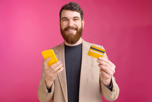 Vidám fiatalember szakállas öltönyben okostelefonnal és hitelkártyával, mobil internetes bankolás - Fotó, kép