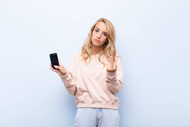 young blonde woman feeling angry, annoyed, rebellious and aggressive, flipping the middle finger, fighting back using a smartphone - 写真・画像