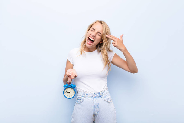 young blonde woman looking unhappy and stressed, suicide gesture making gun sign with hand, pointing to head holding a clock - Foto, imagen