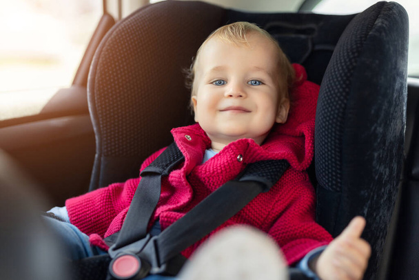 Cute caucasian toodler boy sitting in child safety seat in car during road trip. Adorable baby smiling and enjoying trip in comfortable place in vehicle. Children care and safety on road - Fotó, kép