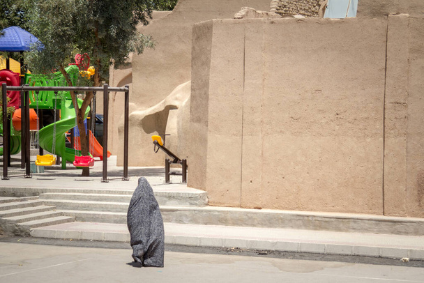 Gestalt einer Frau, die in den Straßen von Yazd, Iran, an einem Kinderspielplatz vorbeigeht und einen Niqab trägt, einen integralen, bescheidenen Kleidungsschleier, der Kopf und Körper bedeckt - Foto, Bild