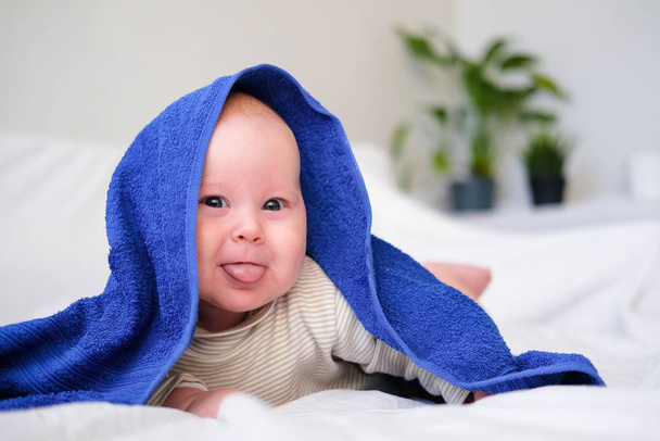 Cute smiling caucasian infant baby looking at camera in blue towel after bathing in the bath on white bed.Copyspace. - Foto, Bild